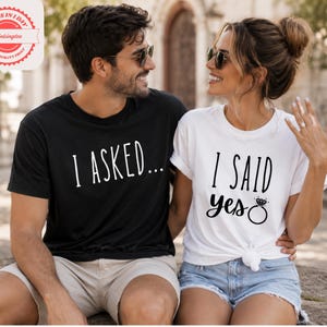 A man and a woman sitting next to each other, smiling and posing for a photo. They are wearing matching t-shirts with the text "I asked... I said yes" printed on them.