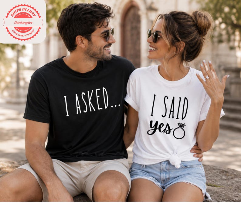 A man and a woman sitting next to each other, smiling and posing for a photo. They are wearing matching t-shirts with the text "I asked... I said yes" printed on them.