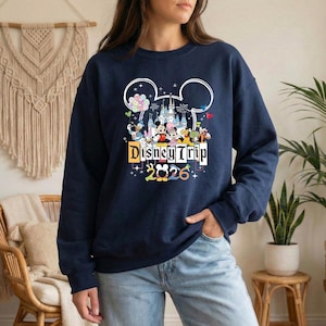 A woman wearing a navy blue sweatshirt with a Disney Trip graphic and text, standing in front of a beige wall with a macrame decoration.