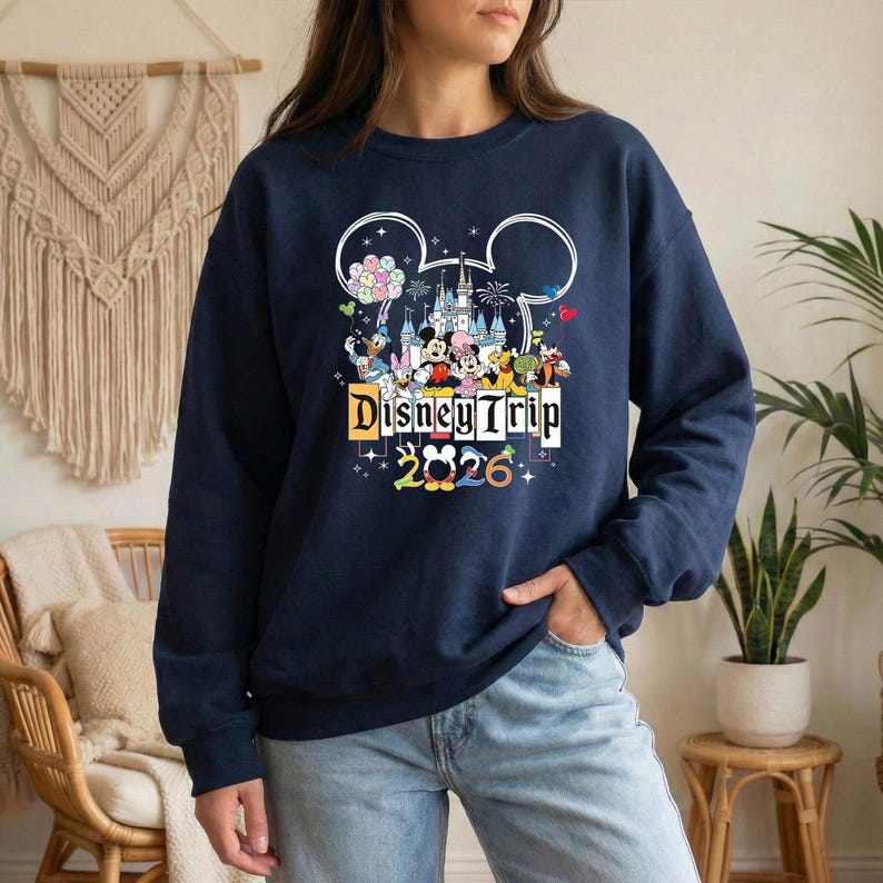 A woman wearing a navy blue sweatshirt with a Disney Trip graphic and text, standing in front of a beige wall with a macrame decoration.