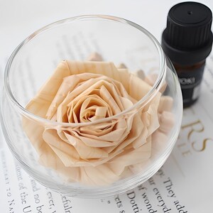 Puede incluir: Un cuenco de vidrio transparente contiene una rosa color melocotón, con sus pétalos dispuestos en espiral. A la derecha, una pequeña botella marrón oscuro con tapa negra. El fondo es una superficie blanca con texto impreso. Tema floral y delicado.