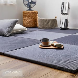 以下が含まれることがあります： 木製の床に敷かれた灰色の畳。畳は編まれたわらでできており、表面はざらざらしています。畳の上にコーヒーカップと小さな木のトレイがあります。