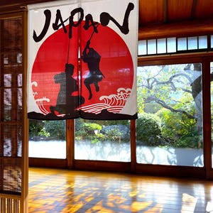 以下が含まれることがあります： 「JAPAN」の文字が黒い筆記体で書かれた、白と赤の日本の暖簾。赤い太陽と2人の黒い忍者の姿、そして波が描かれています。木製の床と大きな窓がある部屋に吊るされています。