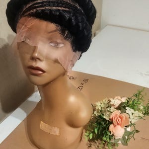May include: A black braided wig with a lace front is displayed on a mannequin head. The mannequin is a light tan color and has a small bouquet of pink and white flowers next to it.