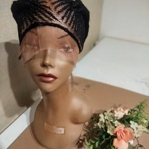 May include: A black braided wig with a lace front is displayed on a mannequin head. The wig features intricate cornrow patterns. A small bouquet of peach and white flowers sits nearby.