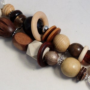 May include: A handcrafted wooden bead necklace featuring a variety of shapes and colors. The beads include spheres, cylinders, and geometric shapes in shades of brown, beige, and cream. The necklace is strung on a silver chain.