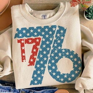 Könnte beinhalten: Ein beiger Pullover mit einer roten und blauen Grafik der Zahl 76 mit Sternen. Die Grafik ist abgenutzt und hat einen Vintage-Look.