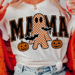 Könnte beinhalten: Weißes T-Shirt mit einem Halloween-Motiv. Das Wort "MAMA" ist in schwarzen Blockbuchstaben mit orangefarbener Umrandung geschrieben. Ein kariertes Gespenst, zwei Fledermäuse und zwei Kürbisse vervollständigen das Design. Das T-Shirt ist an der Taille gebunden.