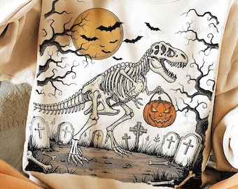 May include: Cream-colored sweatshirt featuring a Halloween-themed graphic. The design includes a skeletal T-Rex holding a jack-o'-lantern, a full moon, bats, and a graveyard scene with tombstones and bare trees. The overall aesthetic is spooky and festive.