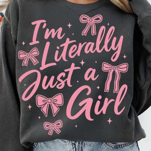 May include: A black sweatshirt with a pink graphic that says "I'm literally just a girl" with pink bows around the text.