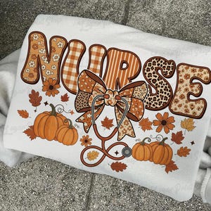 May include: White sweatshirt with a fall-themed design. The word "NURSE" is displayed in a mix of patterns, including floral, plaid, and leopard print. A stethoscope and pumpkins are also featured in the design, along with fall leaves and flowers.