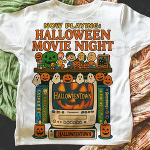 May include: White t-shirt with a Halloween movie night graphic. The design features a retro TV displaying a jack-o'-lantern, surrounded by pumpkins, ghosts, and candles. Text includes "Now Playing: Halloween Movie Night" and "Halloweentown".