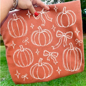 May include: A rust-colored fabric item featuring a white line-art pattern of pumpkins, decorative bows, and four-pointed stars scattered across the surface. The design is printed in a simple, hand-drawn style against the solid orange-brown background.