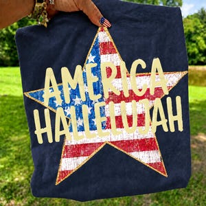 May include: Navy blue t-shirt featuring a large star design in the colors of the American flag. The star is filled with red and white stripes and blue stars. The words "AMERICA HALLELUJAH" are printed in a gold color.