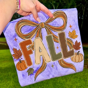 May include: A light purple t-shirt featuring a large graphic of a brown and tan ribbon bow with the word FALL printed in block letters. The design includes orange and brown autumn leaves, small orange pumpkins, and gold star accents.