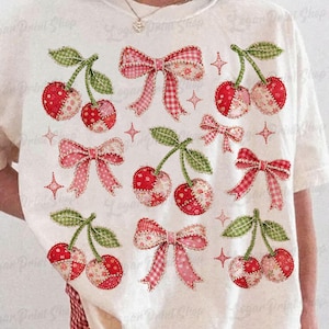 May include: Cream-colored t-shirt featuring a pattern of red cherries and pink bows. The cherries have green leaves and are made of patchwork fabric. The bows are in various styles and patterns, with star accents.