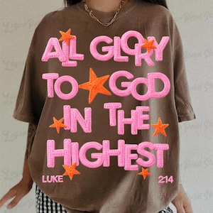 May include: Brown t-shirt with the text "ALL GLORY TO GOD IN THE HIGHEST" in pink, accented with orange starfish. The text includes the reference "LUKE 214".
