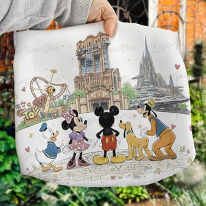 May include: White tote bag featuring a colorful illustration of Disney characters Mickey Mouse, Minnie Mouse, Donald Duck, Goofy, and Pluto. The design includes iconic theme park landmarks and the words "Toy Story" in a playful font. Hearts and floral accents complete the design.