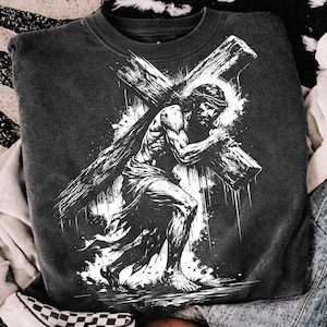 以下が含まれることがあります： 木製の十字架を運ぶ人物の白黒イラストが描かれた、濃いグレーのスウェットシャツ。ハローと茨の冠をかぶり、布をまとった人物が描かれています。デザインは、使い古されたようなヴィンテージ感があります。