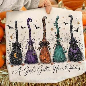 May include: A white t-shirt featuring a graphic of five stylized witch brooms in black, purple, orange, and teal, decorated with stars, moons, bats, and spiderwebs, with the text A Girl's Gotta Have Options printed in black script at the bottom.