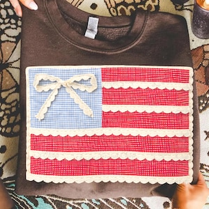 May include: A brown sweatshirt with a sewn-on patch resembling the American flag. The flag design features a blue gingham field with a lace bow, and red and white striped sections with lace trim.
