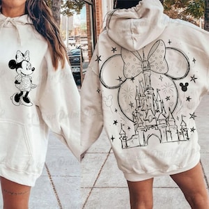 Puede incluir: Sudadera con capucha color crema con un gráfico de Minnie Mouse en blanco y negro en la parte delantera. La parte trasera muestra un gran lazo, orejas de Mickey Mouse, un castillo y un diseño de fuegos artificiales. La sudadera tiene un bolsillo delantero y una capucha con cordones.