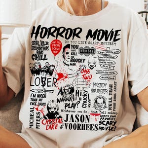 May include: White t-shirt with a graphic design featuring the words "HORROR MOVIE" and various horror movie references, including characters and locations. The design is in black and red.