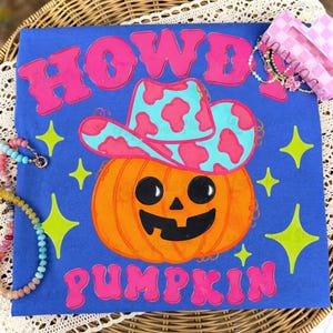 May include: A blue t-shirt featuring a graphic of an orange jack-o'-lantern wearing a light blue and pink cow-print cowboy hat. The text "HOWDY" appears in pink above the pumpkin, and "PUMPKIN" appears in pink below, surrounded by lime green stars.
