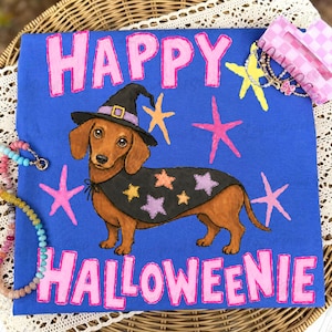 Puede incluir: Una camiseta azul con un perro salchicha marrón que lleva un sombrero de bruja negro y una capa negra decorada con estrellas de colores. El texto "HAPPY HALLOWEENIE" está impreso en grandes letras rosas sobre y bajo el perro, con estrellas.