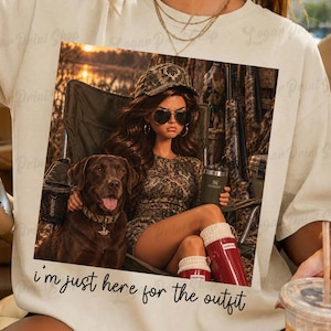 May include: A cream-coloured t-shirt featuring a graphic print of a person wearing a camouflage hat, sunglasses, and a camouflage outfit, sitting in a chair next to a brown Labrador retriever. The text "i'm just here for the outfit" is printed below.