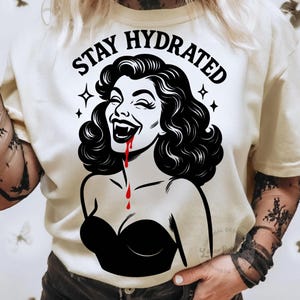 May include: Cream-colored t-shirt with a black and white graphic of a laughing woman with fangs and dripping blood. The text "STAY HYDRATED" is above the graphic. The woman is wearing a black strapless top.