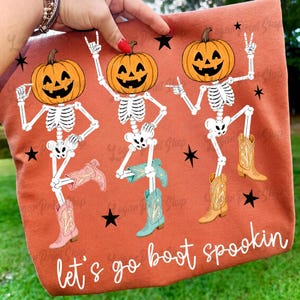 May include: A burnt orange t-shirt featuring a graphic of three skeletons with pumpkin heads wearing colorful cowboy boots. The skeletons are in dancing poses, surrounded by black stars. The text "let's go boot spookin" is printed in white.