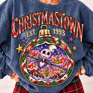 May include: A blue acid-wash sweatshirt featuring a graphic of a Christmas ornament containing a scene with a skeleton character, snowy houses, and trees. The text reads CHRISTMASTOWN EST. 1993 in red and gold lettering with stars surrounding the ornament.