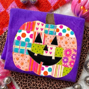 May include: A purple blanket featuring a large central pumpkin graphic composed of various colorful patchwork squares. The pumpkin has a black jack-o'-lantern face with triangular eyes and a jagged mouth, surrounded by patterns of polka dots, stripes, hearts, and flowers.