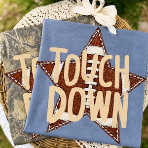 May include: A blue t-shirt featuring a brown star graphic with a football texture and white stitching, overlaid with the beige text "TOUCH DOWN". A second t-shirt with a camouflage pattern is visible underneath, both topped with a white ribbon bow.