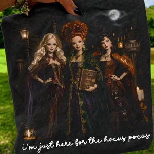 May include: A black t-shirt featuring a graphic print of three dolls dressed in elaborate gowns, one holding a book labeled Book of Spells. The background shows a night scene with a full moon and a sign reading Salem 1692. Text reads i just here for the hocus pocus.