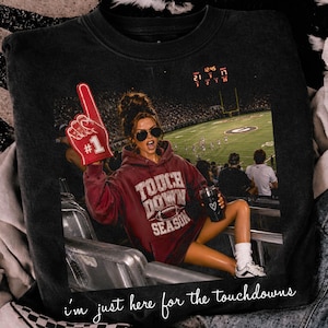 May include: A black crewneck sweatshirt featuring a graphic print of a person in a maroon hoodie holding a red foam finger at a football stadium. The graphic includes the text "TOUCH DOWN SEASON" and "i'm just here for the touchdowns".
