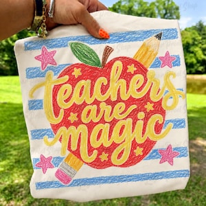 May include: A white fabric tote bag featuring a red apple graphic with a green leaf and a yellow pencil. The text "teachers are magic" is printed in yellow cursive script across the apple. The background has blue horizontal stripes and pink stars.