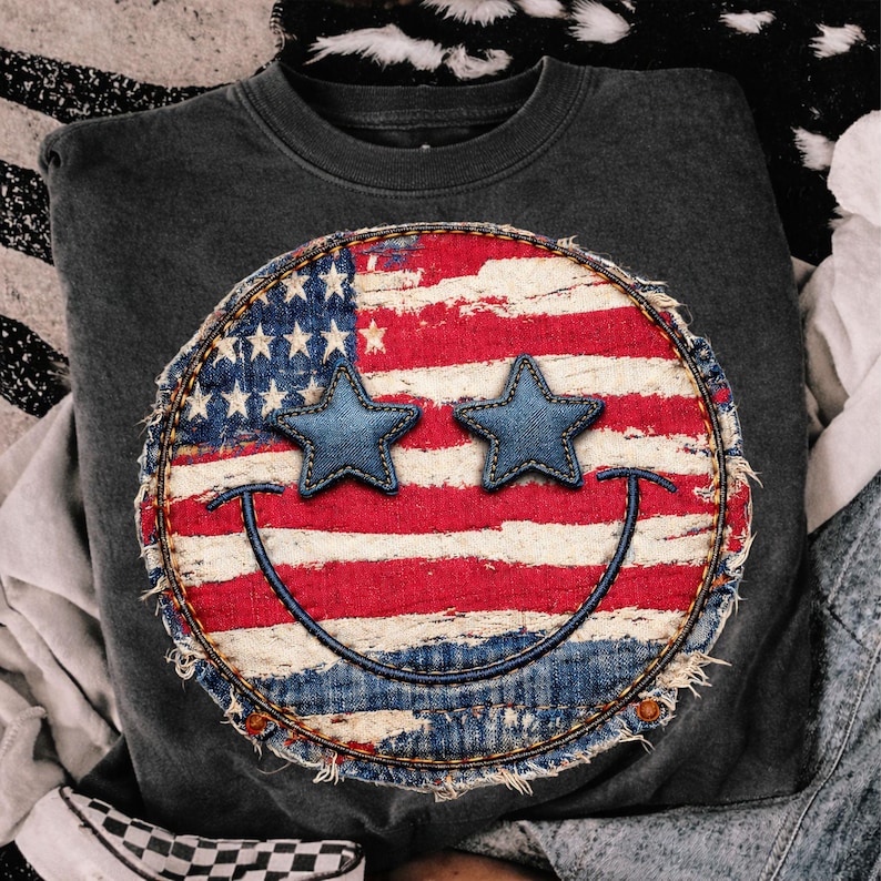 May include: Dark gray sweatshirt featuring a distressed circular design with the American flag and two blue star eyes. The design includes a stitched denim border and a stitched smile. The sweatshirt is a casual style.