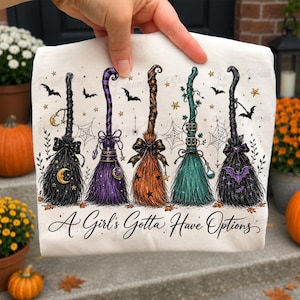 May include: A white t-shirt featuring a graphic of five distinct witch brooms in black, purple, orange, teal, and black, decorated with stars, spiderwebs, bats, and moons. The text A Girl's Gotta Have Options is printed in black script below.