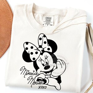 May include: A white t-shirt with a black and white image of Minnie Mouse. The image shows Minnie Mouse with her signature polka dot bow and a playful expression. The text "Minnie Mouse xoxo" is printed below the image.