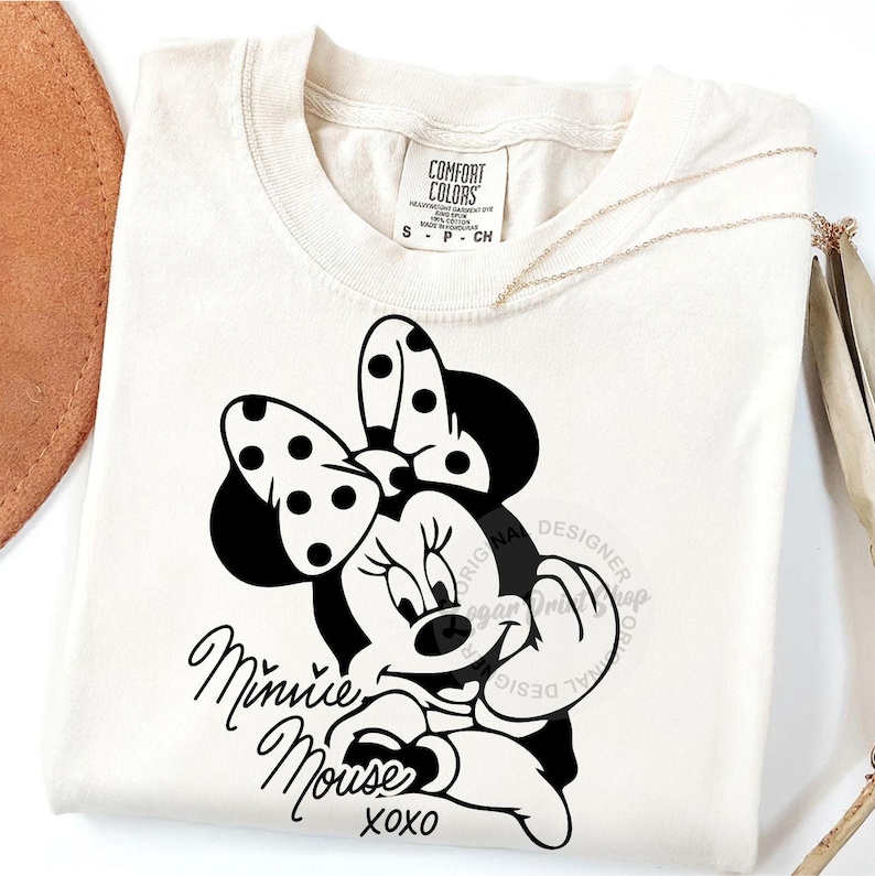 May include: A white t-shirt with a black and white image of Minnie Mouse. The image shows Minnie Mouse with her signature polka dot bow and a playful expression. The text "Minnie Mouse xoxo" is printed below the image.