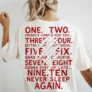 May include: Cream-colored t-shirt with red text and claw marks. The text reads "ONE, TWO, FREDDY'S COMING FOR YOU... THREE FOUR, BETTER LOCK YOUR DOOR..." and continues with the numbers five through ten. A horror-themed graphic tee.