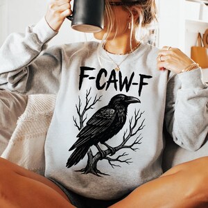 May include: A light gray sweatshirt featuring a black raven perched on a tree branch. The text "F-CAW-F" is printed above the raven. The sweatshirt has long sleeves and a crew neck.