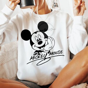 May include: White sweatshirt with a black and white image of Mickey Mouse. The image includes the text "Mickey Mouse" in a cursive font.