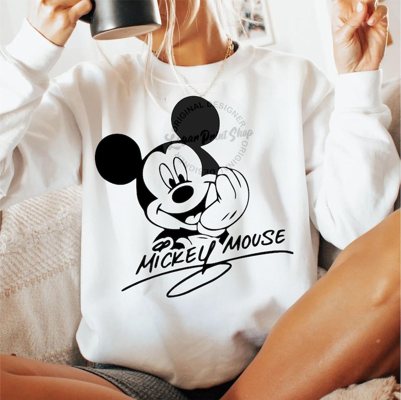 May include: White sweatshirt with a black and white image of Mickey Mouse. The image includes the text "Mickey Mouse" in a cursive font.
