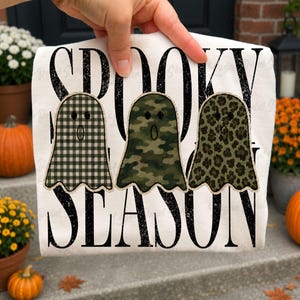 May include: A white t-shirt featuring the black text SPOOKY SEASON and three ghost illustrations. The first ghost has a black and white buffalo plaid pattern, the second has a green camouflage print, and the third has a green leopard print.