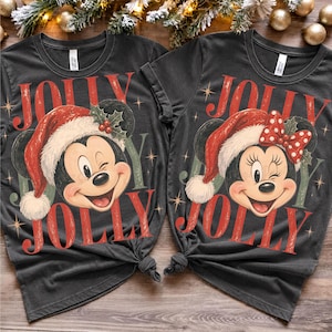 May include: Two dark gray t-shirts featuring a graphic of Mickey Mouse and Minnie Mouse wearing red Santa hats with holly accents. The text JOLLY is printed in large red letters above and below the characters, surrounded by small star icons.