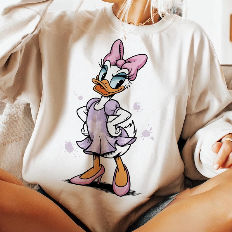 May include: White sweatshirt with a cartoon image of a duck wearing a purple dress and a pink bow. The duck is standing on a white background with purple watercolor splatters.