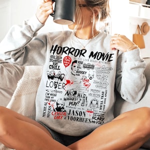 May include: A light gray sweatshirt with a graphic design featuring the words "HORROR MOVIE" and various horror movie references, including characters and phrases. The design is in black and red.